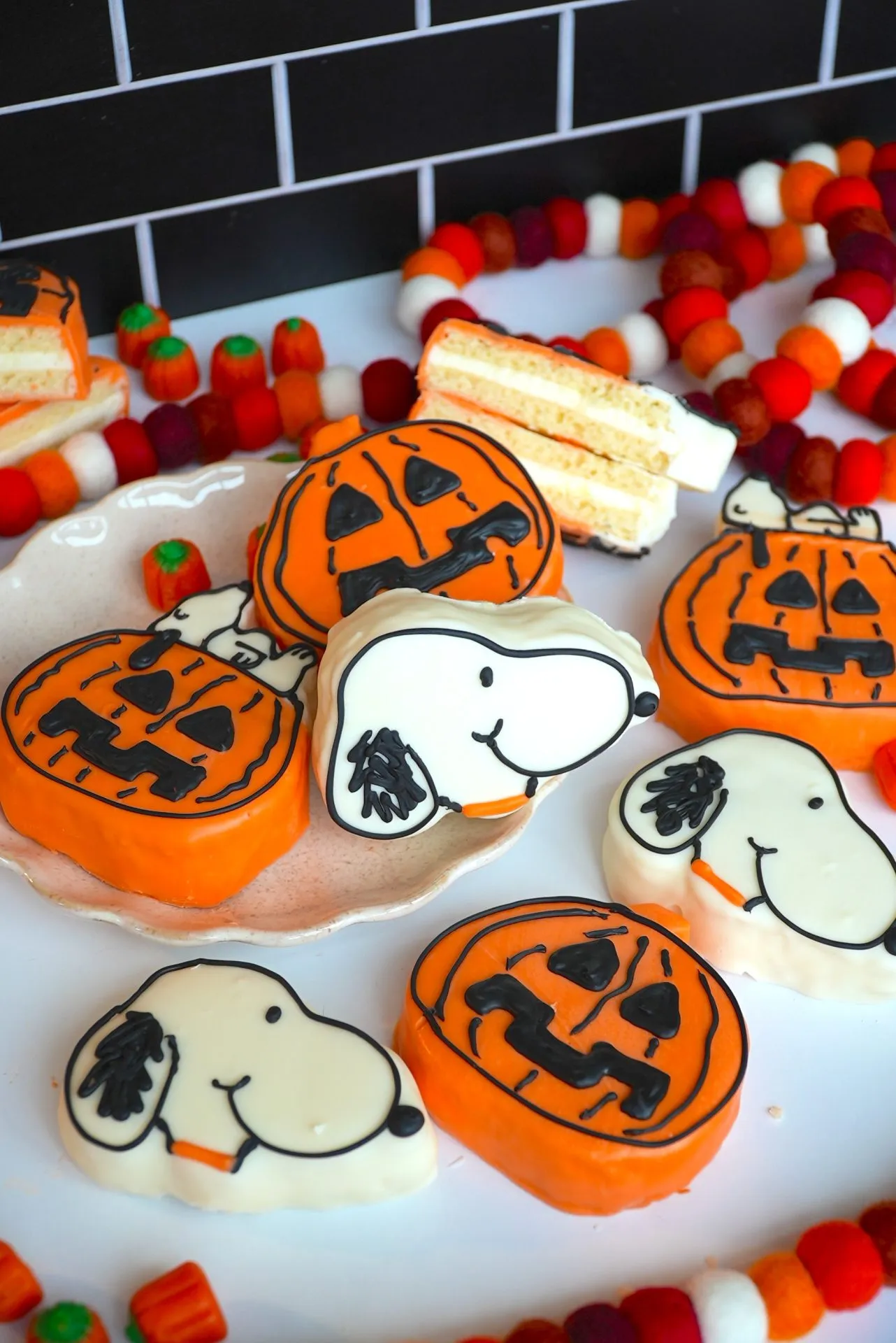Halloween-themed cookies shaped like pumpkins with jack-o'-lantern faces and Snoopy's head are displayed on a plate, surrounded by festive decorations and treats on a white surface.