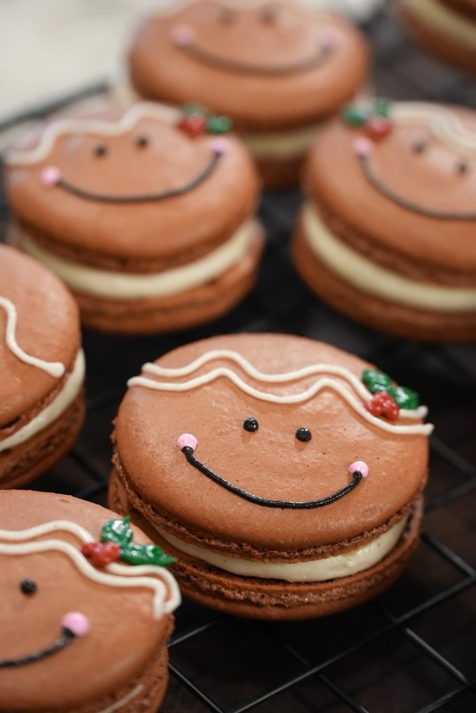 Chocolate macarons decorated with smiling gingerbread faces, pink cheeks, white icing hair, and small holly decorations, arranged on a black cooling rack.