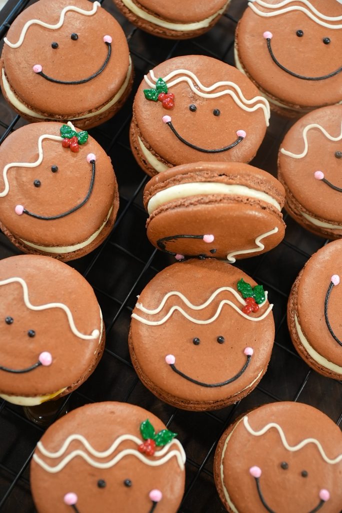 Gingerbread-style macarons with smiling faces, pink cheeks, white icing details, and small holly decorations, arranged on a black cooling rack.