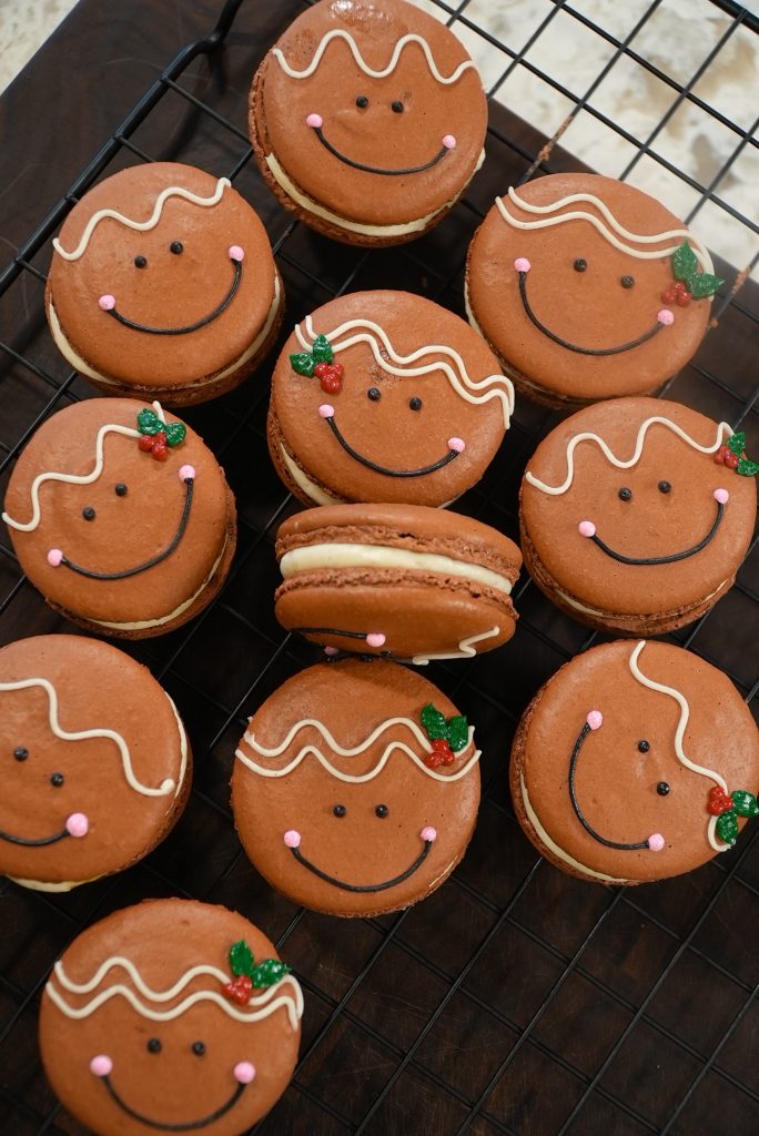 A batch of brown macarons decorated with smiling gingerbread faces, white icing squiggles, pink cheeks, and small green and red holly accents, displayed on a cooling rack.
