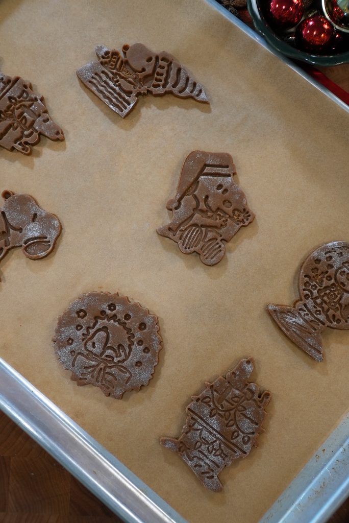 A baking tray lined with parchment paper holds six unbaked chocolate cookies shaped like Peanuts characters and festive holiday items, ready to be baked. Ornaments can be seen in the corner of the image.