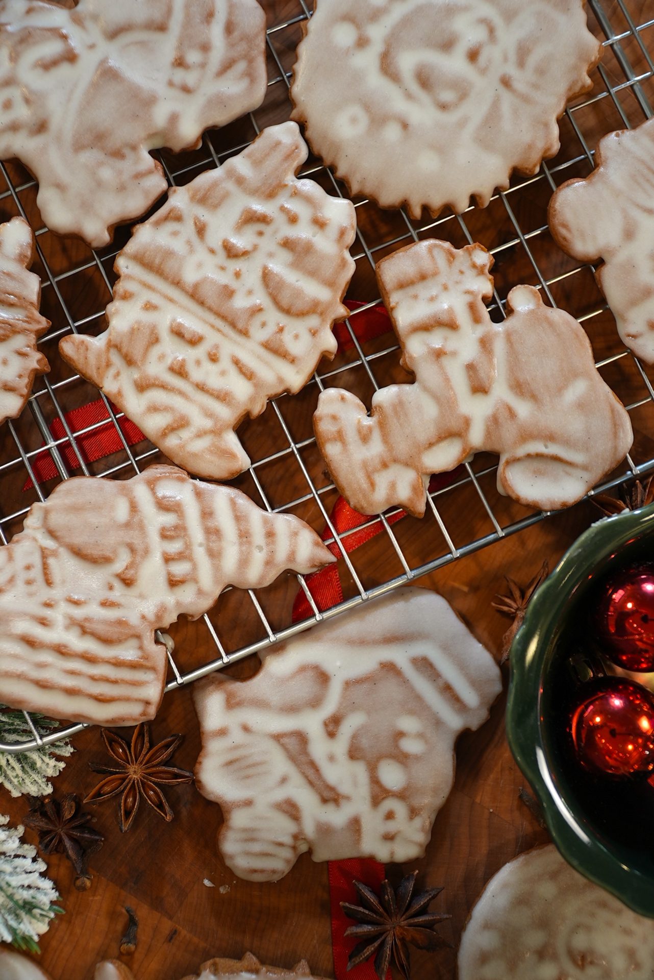 Iced Christmas cookies in festive shapes cool on a wire rack. A green bowl with red ornaments and some star anise are visible nearby on a wooden surface decorated for the holidays.