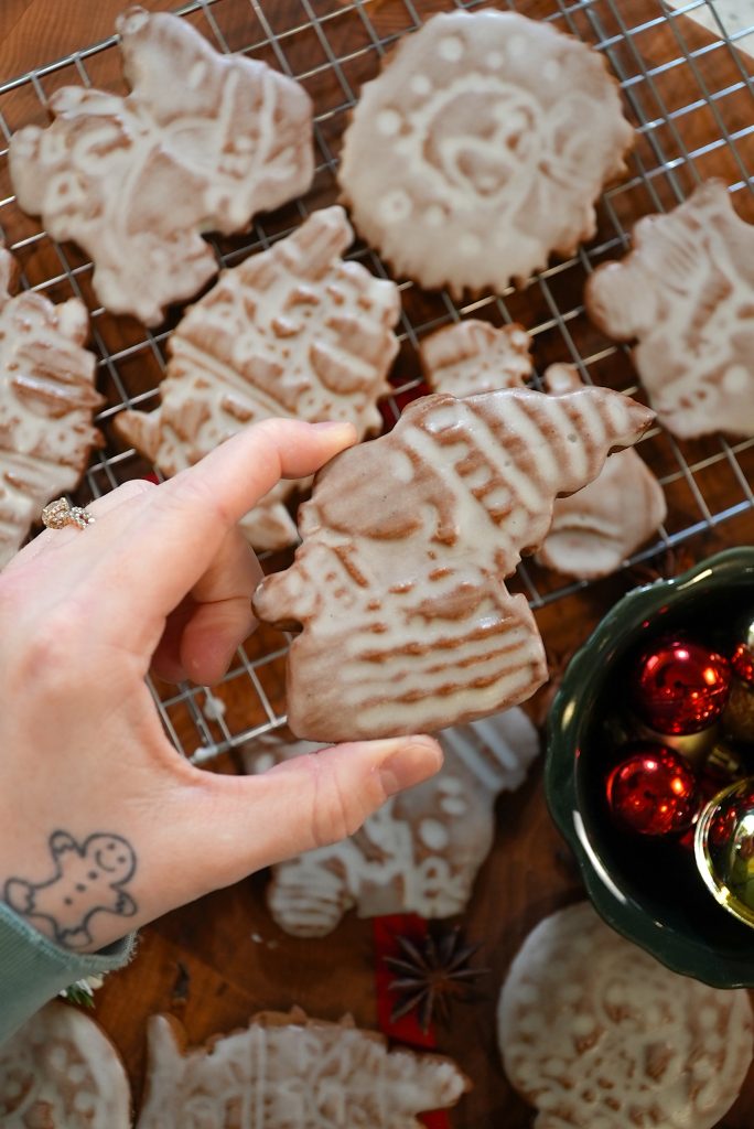 A hand holding a decorated, iced Christmas cookie shaped like a gnome. More iced cookies and a cooling rack are in the background, with a small bowl of red Christmas ornaments nearby.