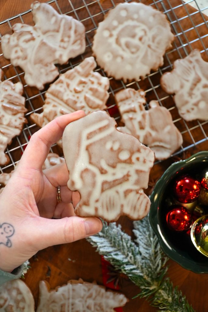 A hand holds a decorated holiday cookie with white icing, shaped like a cartoon character. More iced cookies cool on a wire rack. Nearby are Christmas ornaments in a bowl and a frosted pine branch on a wooden surface.