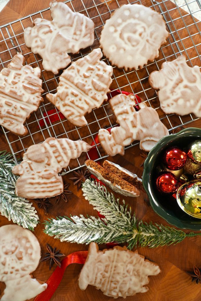 Iced Christmas cookies in festive shapes rest on a cooling rack and wooden surface, with holiday ornaments, fir branches, and star anise nearby, creating a cheerful holiday scene.