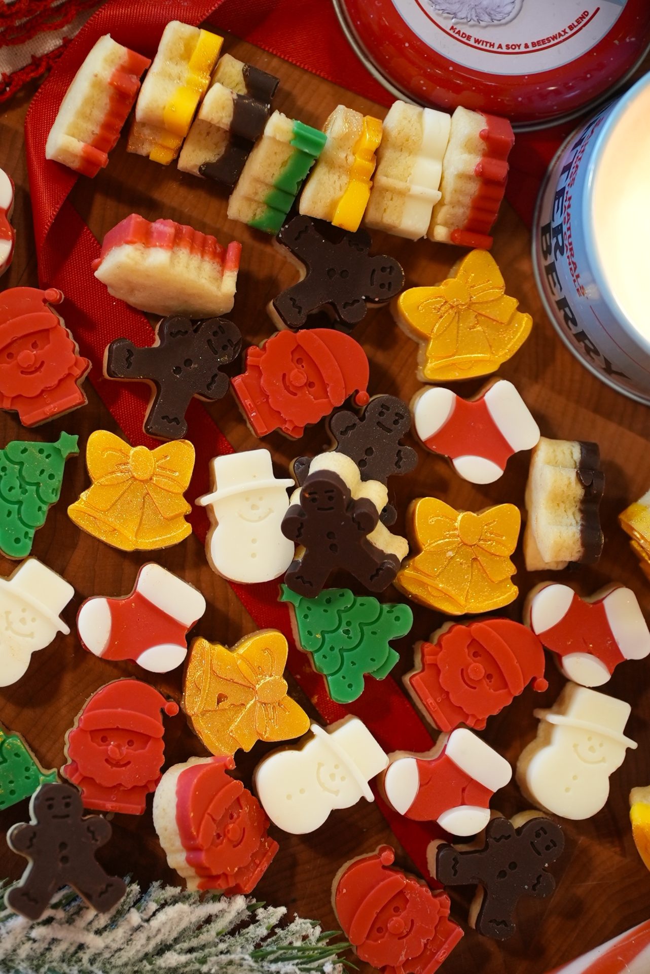 Assorted Christmas-themed soaps in shapes like Santa, snowmen, bells, trees, and gingerbread men, arranged on a wooden surface with a lit candle and festive ribbon nearby.