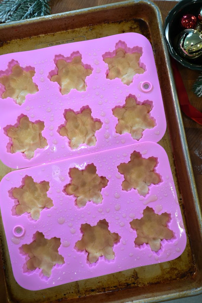 A pink silicone mold with maple leaf shapes is filled with light brown dough and placed on a baking tray. The mold appears to be lightly sprayed with oil. Holiday decorations are partially visible nearby.