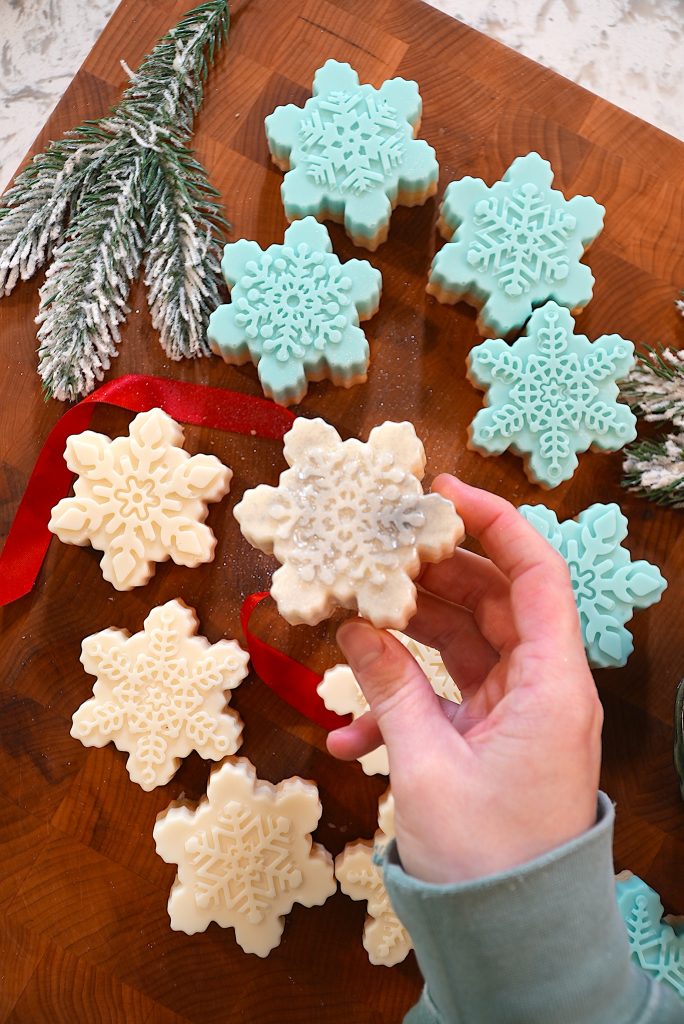 A hand holds a white snowflake-shaped cookie above a wooden board with more snowflake cookies in white and light blue, decorated with intricate icing patterns. Frosted pine branches and a red ribbon are also visible.