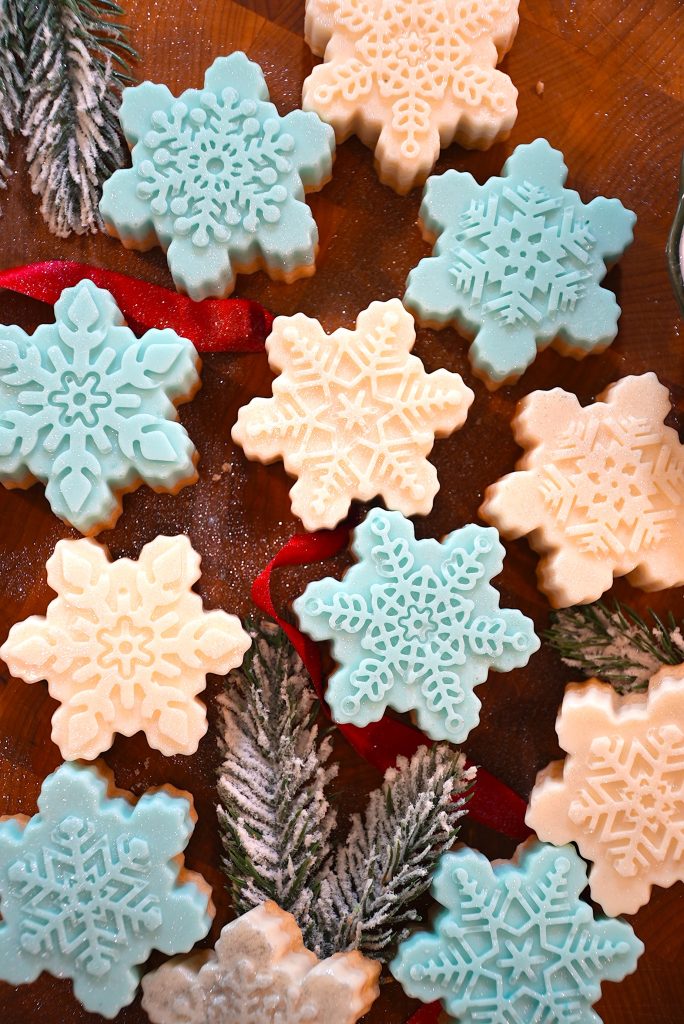 Snowflake-shaped cookies in blue and white are arranged on a wooden surface with frosted pine branches and a red ribbon, creating a festive holiday scene.