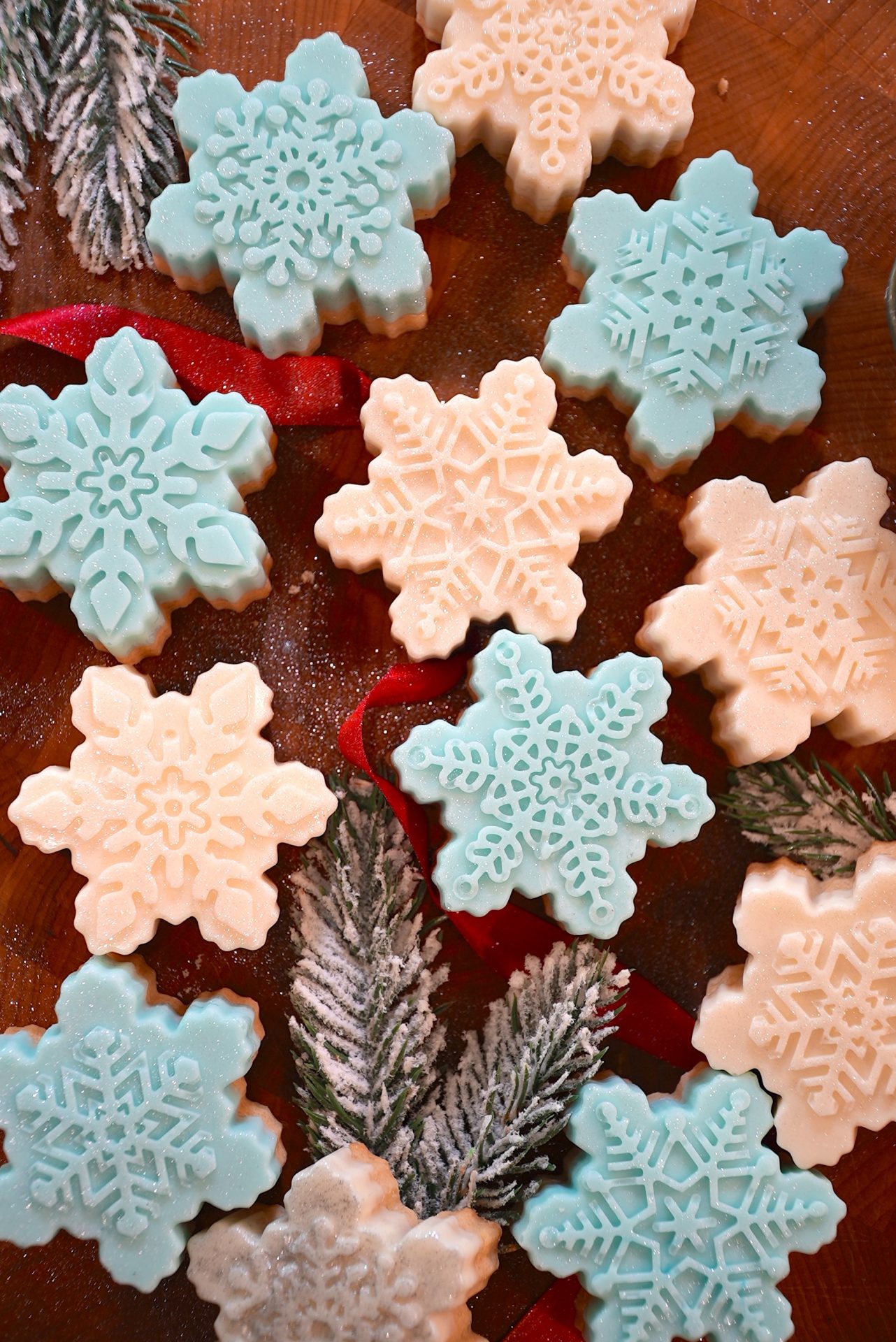 Snowflake-shaped cookies in white and light blue are arranged on a wooden surface, with red ribbon and pine branches dusted with powdered sugar as decoration.