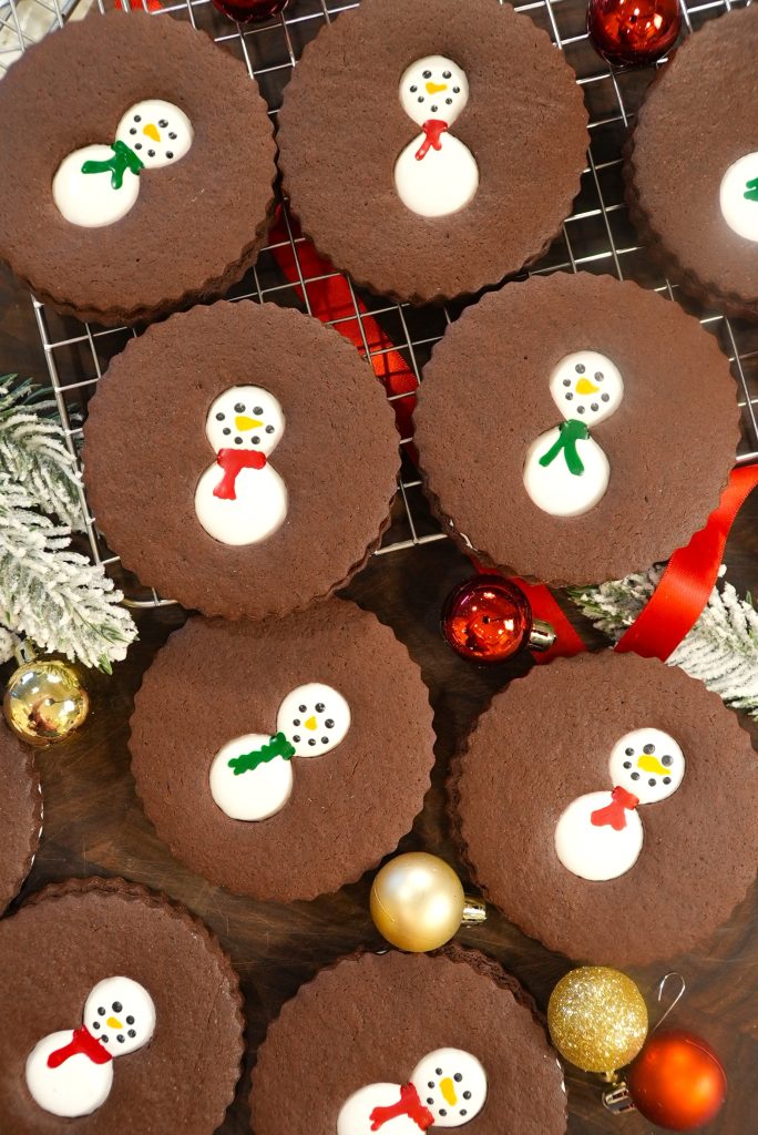 Chocolate cookies with snowman designs in white icing are arranged on a cooling rack and wooden surface, surrounded by red and gold Christmas ornaments and frosted greenery.