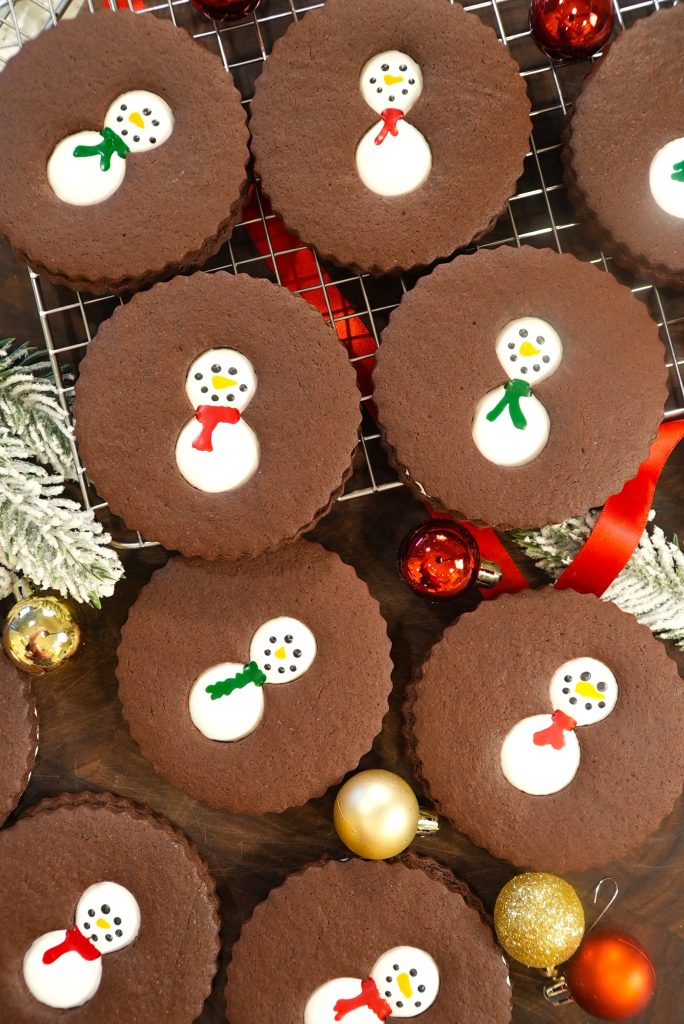 Chocolate cookies with snowman designs, decorated with white icing and colored scarves, arranged on a cooling rack with festive holiday ornaments and frosted greenery nearby.