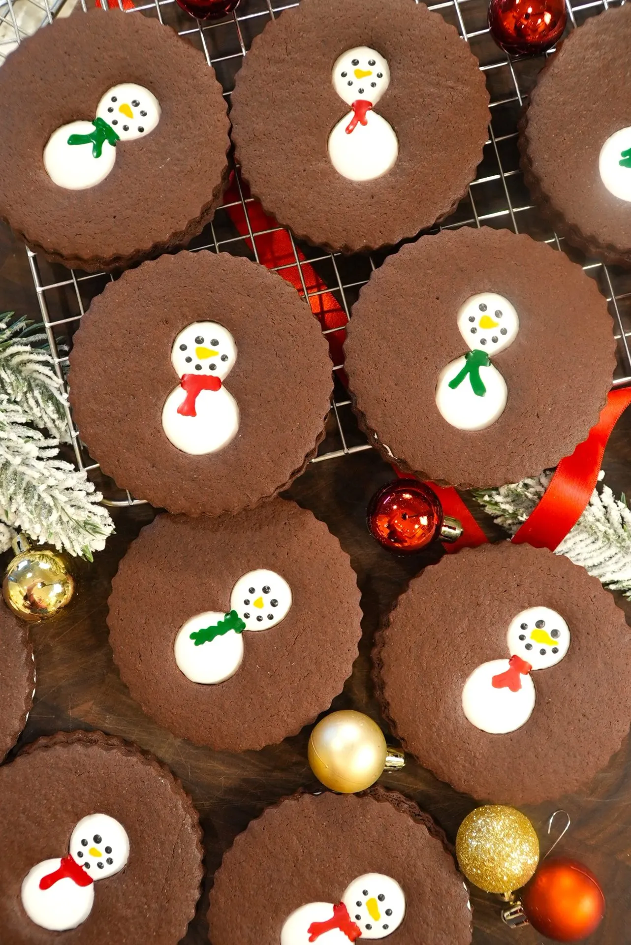 Chocolate cookies with snowman designs, decorated with white icing and colored scarves, arranged on a cooling rack with festive holiday ornaments and frosted greenery nearby.