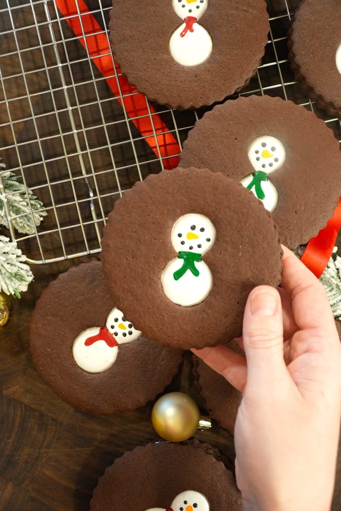 A hand holds a round chocolate cookie with a snowman decoration in the center. More similar cookies are on a cooling rack and wooden surface, with festive ornaments and ribbons nearby.