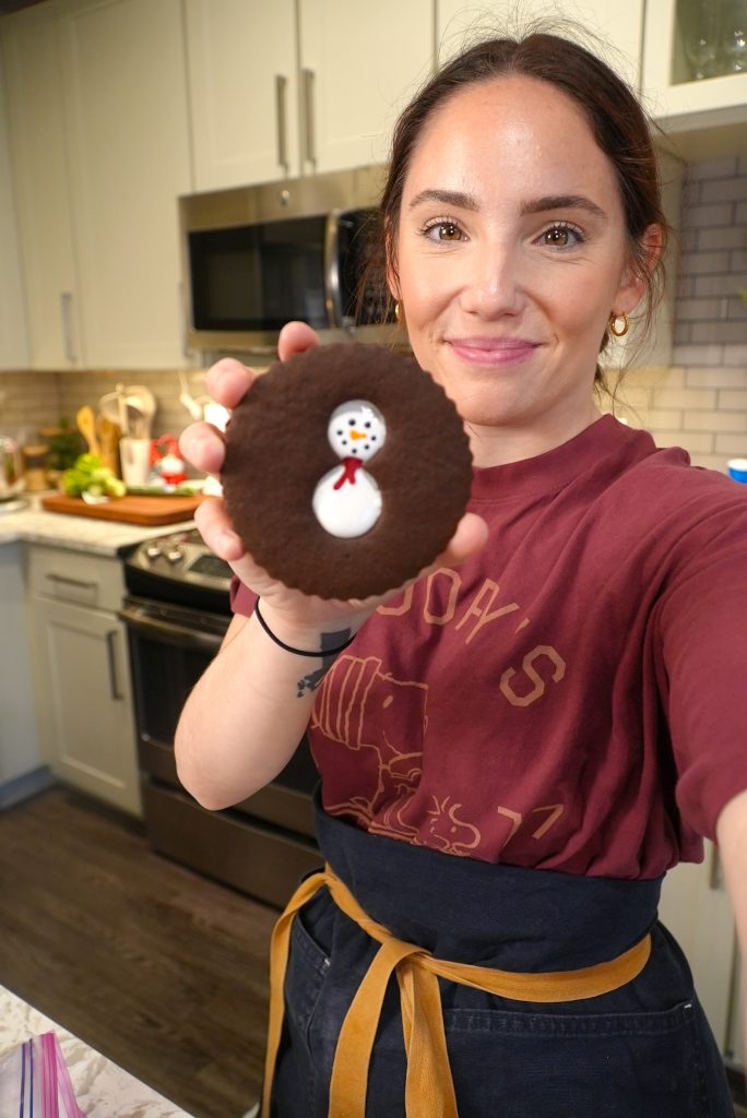 A woman in a maroon shirt and apron holds up a round chocolate cookie decorated with a snowman face, standing in a modern kitchen.