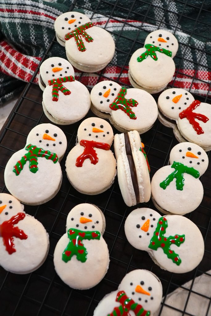 Snowman-shaped macarons decorated with red and green icing scarves, orange carrot noses, and chocolate filling, displayed on a cooling rack with a plaid cloth in the background.