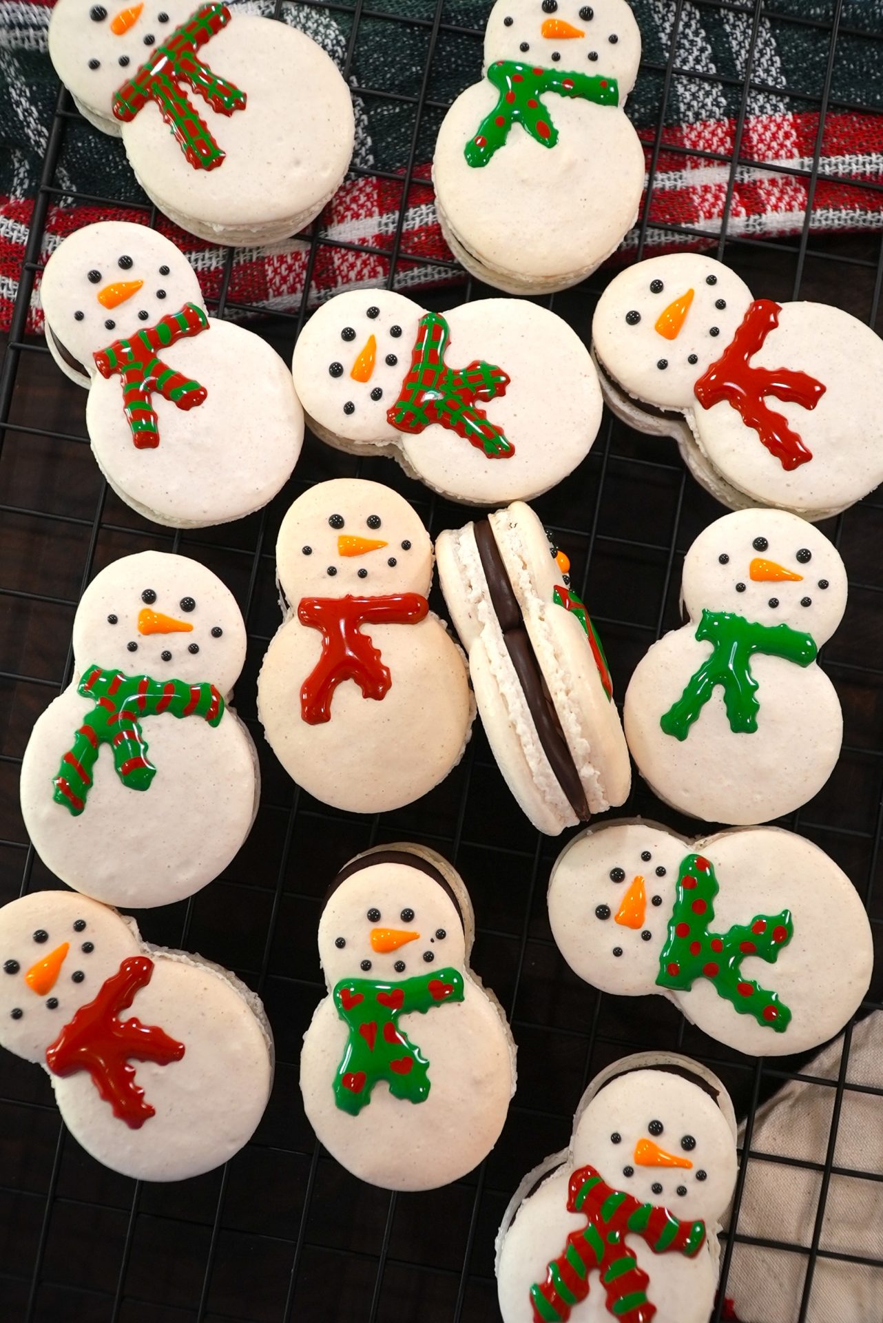 Snowman-shaped cookies with colorful scarves are arranged on a cooling rack. The cookies are decorated with white icing, dotted eyes, orange carrot noses, and either red or green scarves.