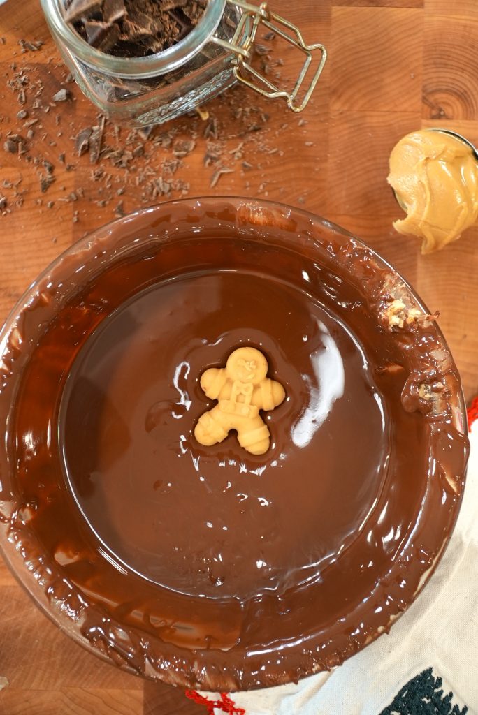 A gingerbread-shaped cookie is partially submerged in a bowl of melted chocolate on a wooden surface, with a jar of chocolate shavings and a spoonful of peanut butter nearby.