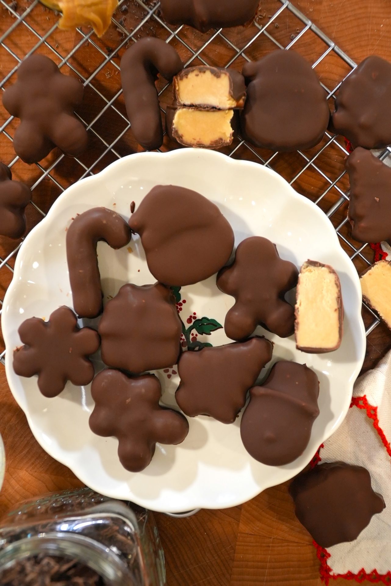 Assorted chocolate-covered, holiday-shaped candies, some cut open to reveal a creamy filling, are arranged on a white plate and cooling rack. Shapes include flowers, gingerbread people, a mitten, and a candy cane.
