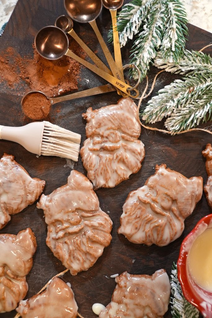 Santa-shaped cookies with a white glaze are arranged on a wooden board. Nearby are frosted pine branches, a brush, a set of measuring spoons, cocoa powder, and a red bowl with more glaze.