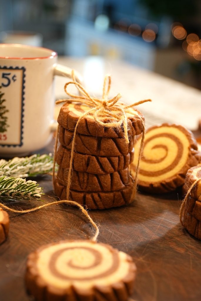 A stack of spiral-patterned cookies tied with twine sits on a wooden surface, with a festive mug, evergreen sprigs, and more cookies in the background. Warm lighting creates a cozy, holiday atmosphere.