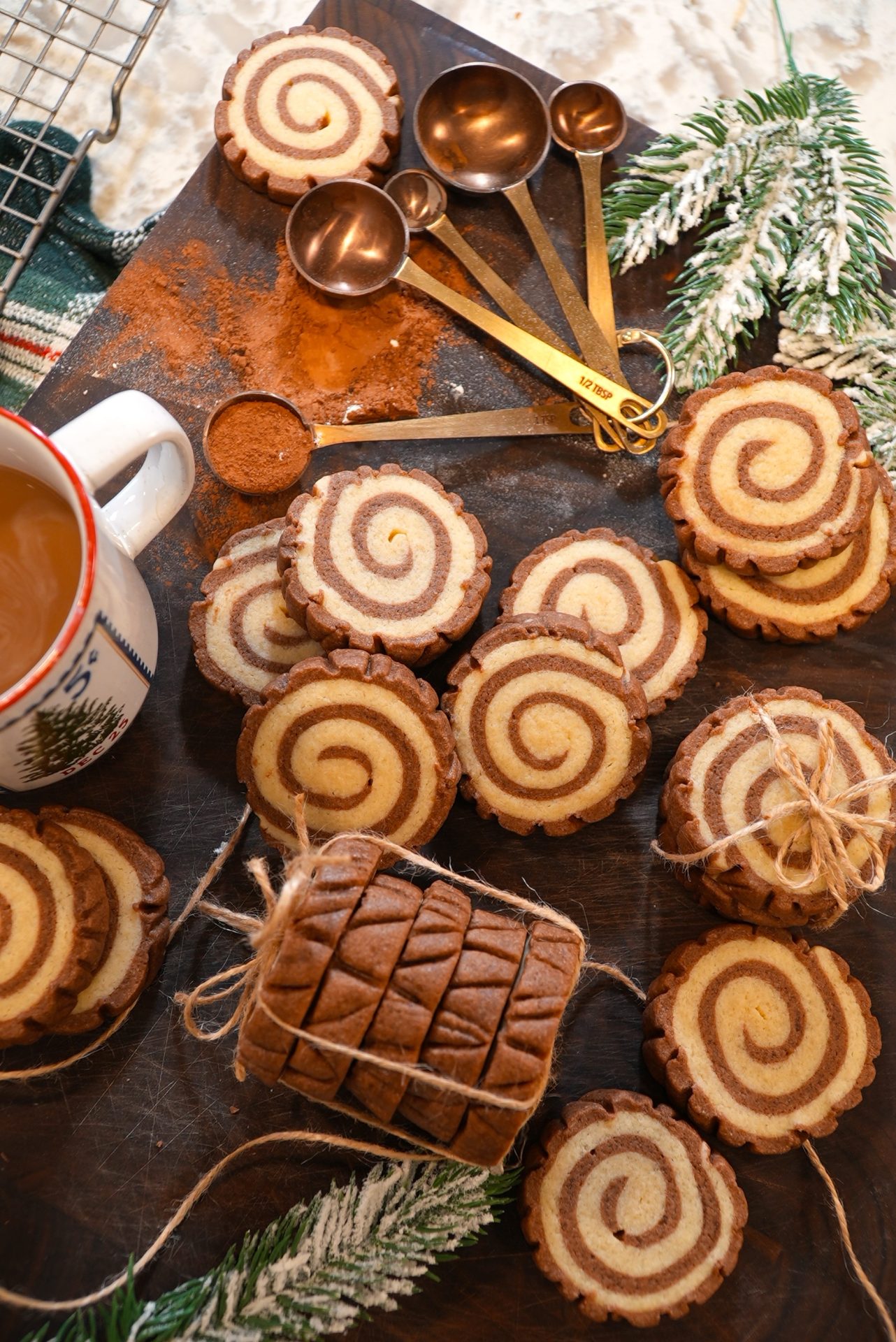 A wooden board with chocolate and vanilla swirl cookies, some tied with string, beside a cup of coffee, copper measuring spoons, cinnamon, and festive pine branches.