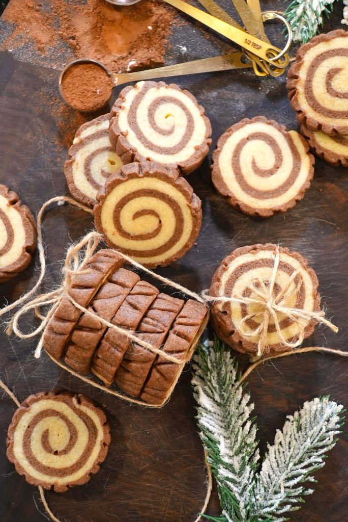 A bundle of chocolate and vanilla pinwheel cookies tied with twine is on a dark wooden surface, surrounded by more cookies, gold measuring spoons, cocoa powder, and frosted pine branches.