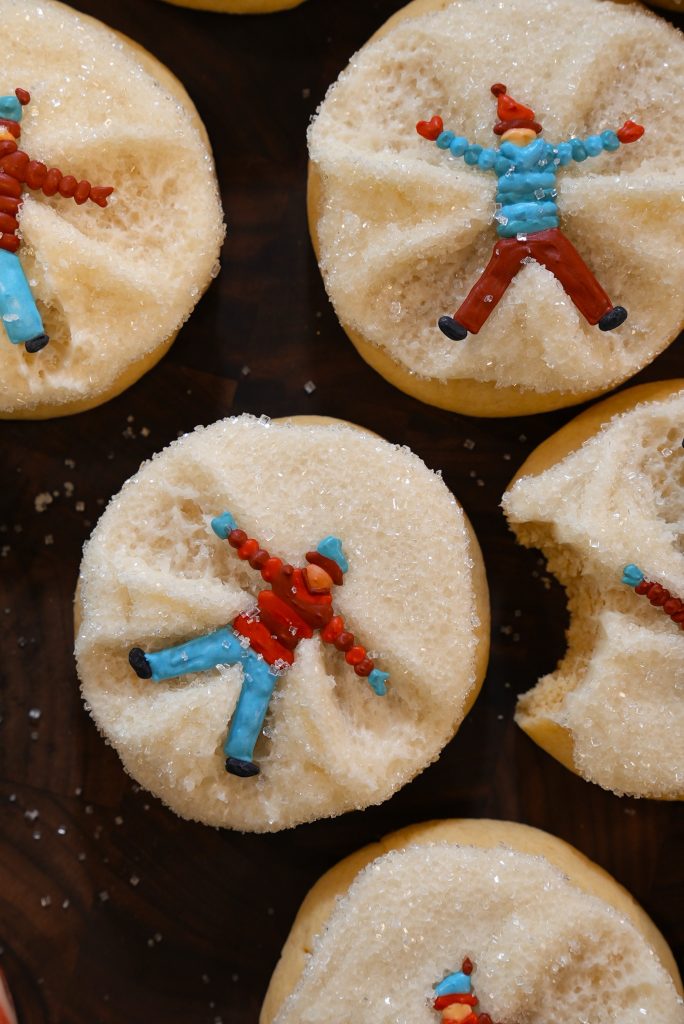 Round sugar cookies decorated with colorful icing figures making snow angels, one cookie has a bite taken out of it. The cookies are arranged on a dark surface.