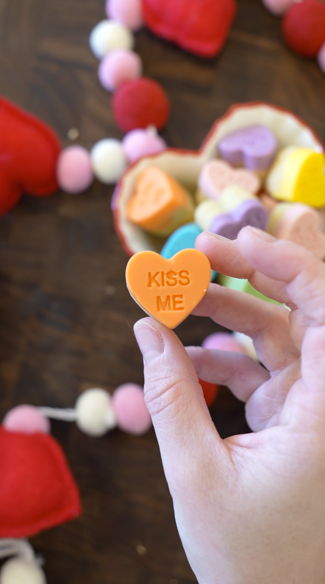 A hand holds an orange heart-shaped candy with "KISS ME" written on it. In the background, there are more colorful heart candies in a bowl and a garland made of red, pink, and white felt balls.