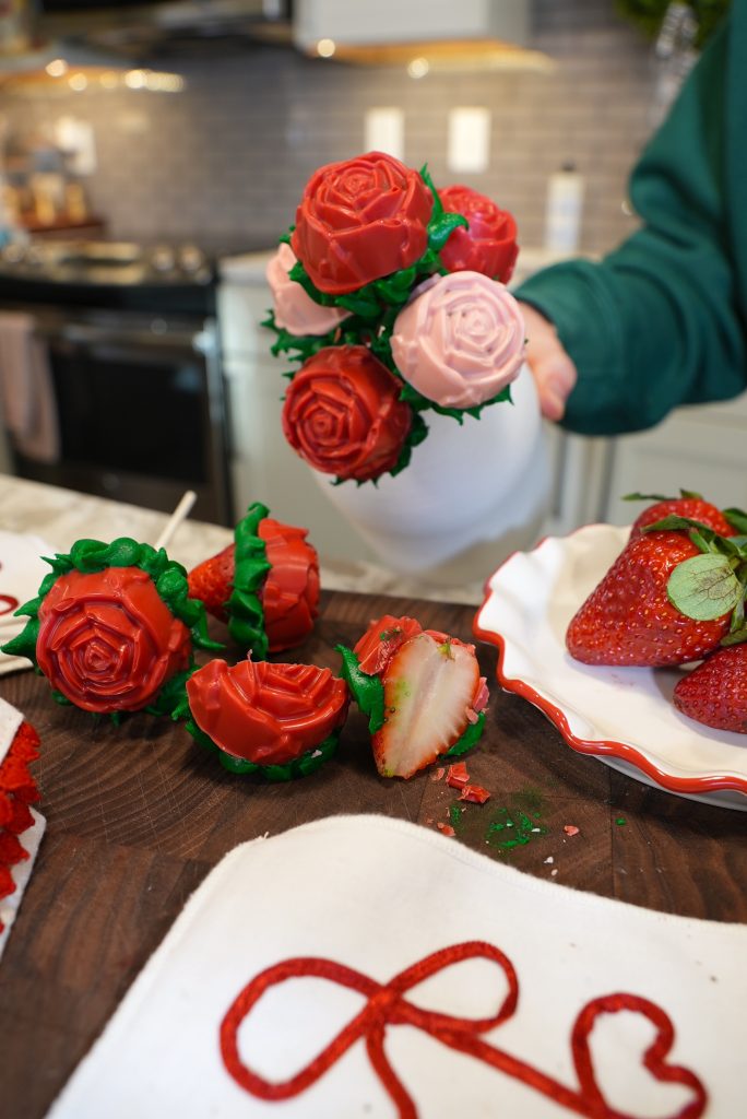 A hand holds a white vase filled with red and pink chocolate-covered strawberries shaped like roses. A plate with strawberries and a cut strawberry rose are on a kitchen counter in the background.