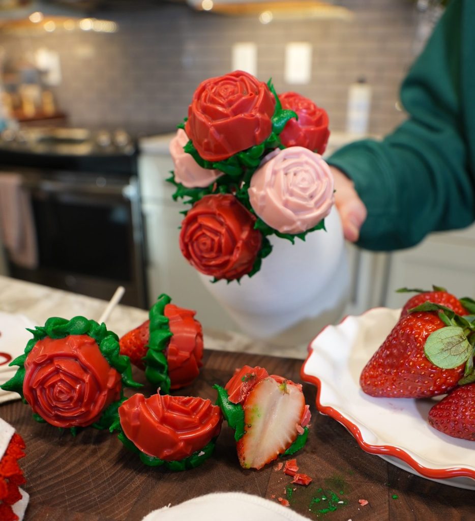 A hand holds a white vase filled with red and pink chocolate-covered strawberries shaped like roses. A plate with strawberries and a cut strawberry rose are on a kitchen counter in the background.