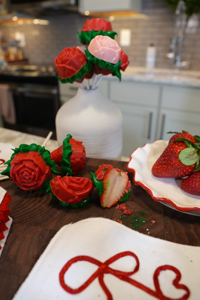 Strawberries decorated to look like red and pink roses are arranged in a white vase and on a table. A cut strawberry shows its interior. A plate with strawberries and a napkin with red embroidery are nearby.