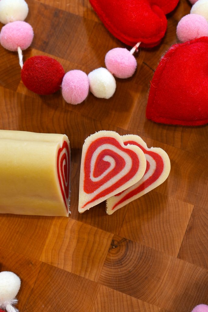 Heart-shaped slices of red and white spiral cookie dough are displayed on a wooden surface, with red felt hearts and pink-and-white pom-pom garland as decoration.