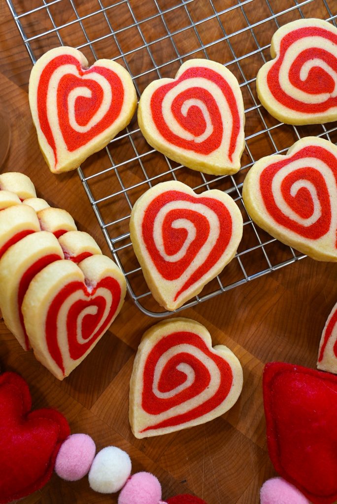 Heart-shaped sugar cookies with red swirls are cooling on a metal rack and wooden surface. Some cookies are stacked, and plush red and white heart decorations are nearby.
