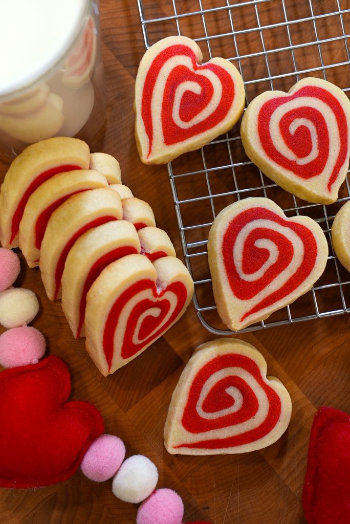 Heart-shaped cookies with red and white swirls are arranged on a wooden surface and a cooling rack. Nearby, there are felt heart decorations and a cup of milk partially visible in the corner.