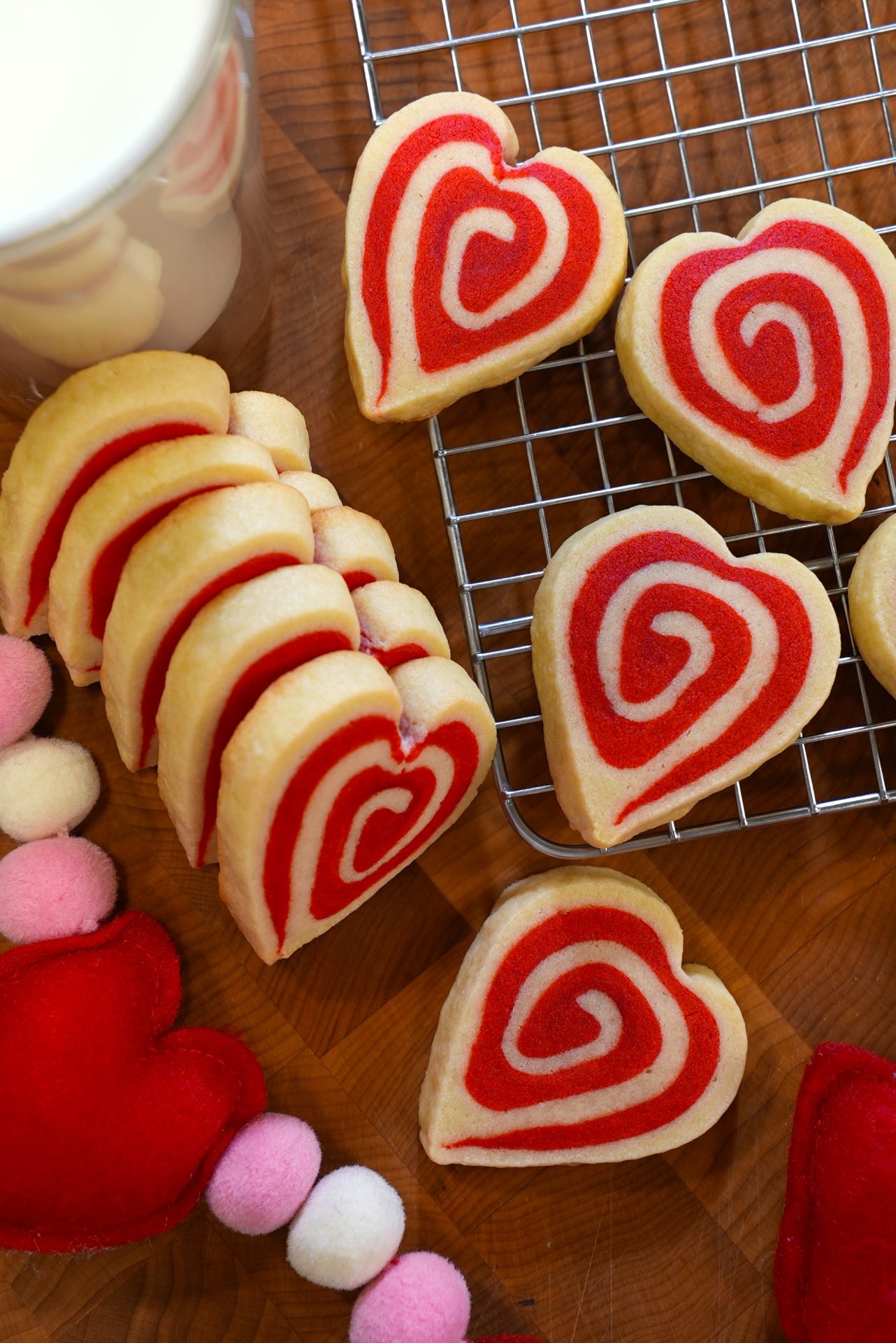 Heart-shaped cookies with red and white swirls are arranged on a wooden surface and a cooling rack. Nearby, there are felt heart decorations and a cup of milk partially visible in the corner.
