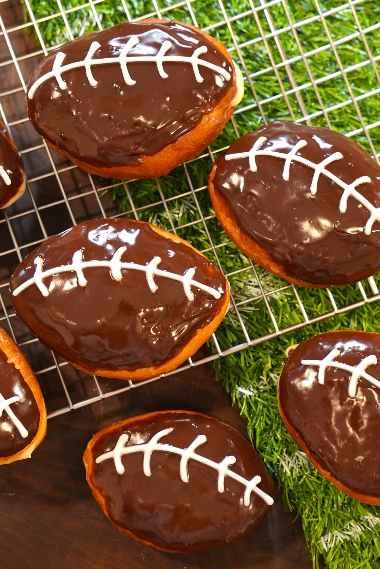 Chocolate-glazed football-shaped donuts with white icing laces sit on a wire rack and artificial grass, resembling footballs, perfect for a sports-themed party or event.