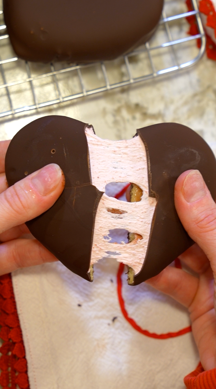 Two hands pull apart a heart-shaped chocolate-covered treat, revealing a stretchy, fluffy pink filling inside. A cooling rack and a red-and-white towel are visible in the background.