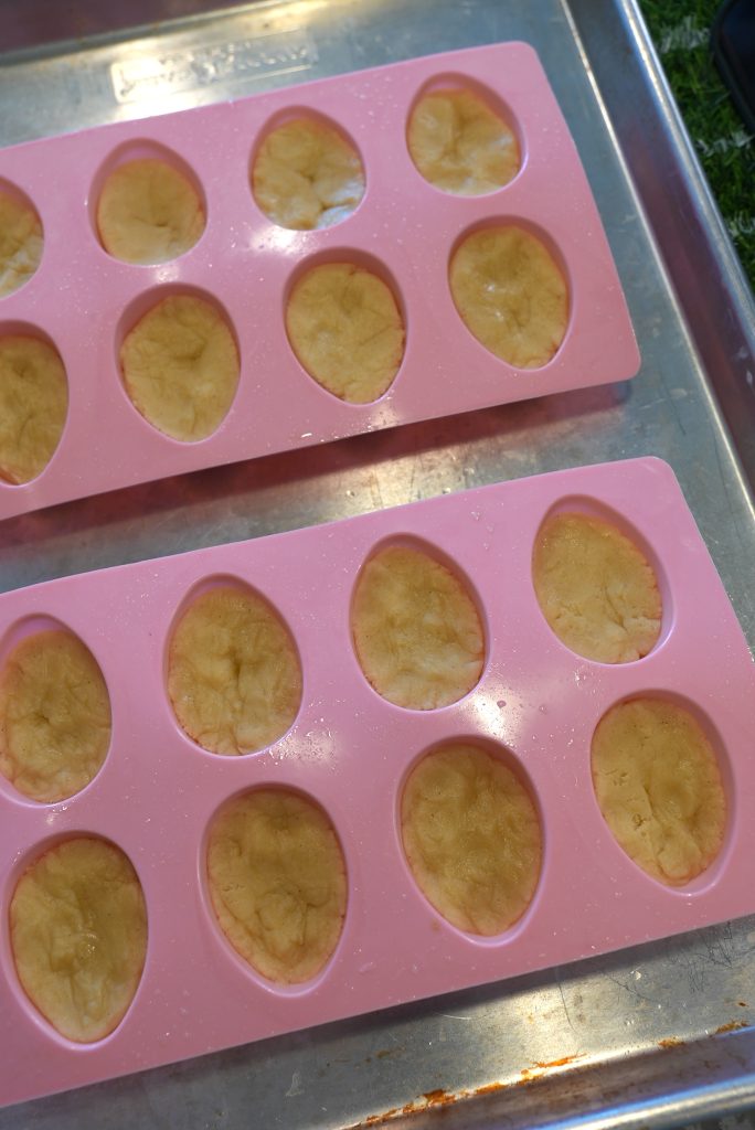 Two pink silicone molds with oval-shaped cavities sit on a metal baking tray. Each cavity is filled with a yellowish dough or batter, ready to be baked or set.