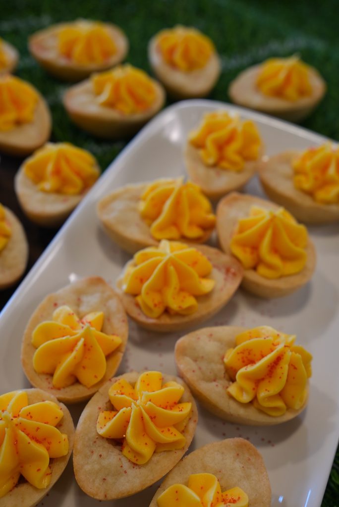A close-up of bite-sized tartlets with golden yellow piped cream on top, arranged on a white platter and in the background, more tartlets are displayed on a green surface.