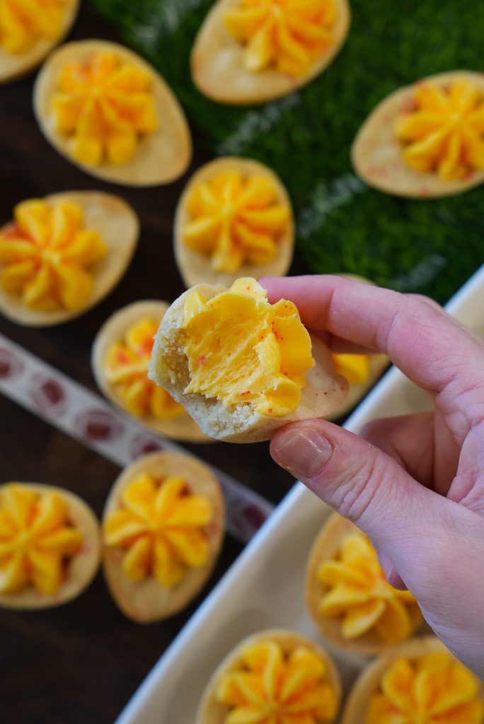 A hand holds a bitten deviled egg-shaped cookie with yellow frosting, above a tray filled with similar cookies decorated to look like deviled eggs.