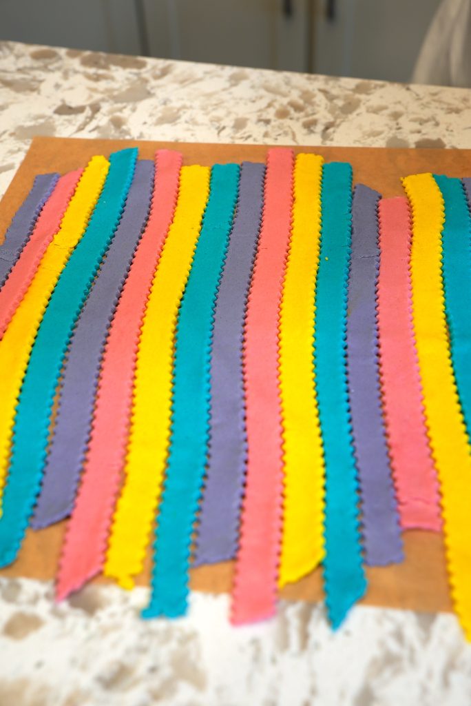 Colorful strips of pasta dough in blue, pink, yellow, and purple are laid side by side on a marble countertop, with zigzag edges visible on each strip.