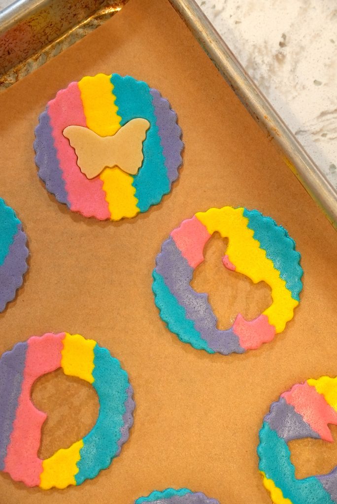 Colorful, round cookies with scalloped edges and striped yellow, pink, blue, and purple dough, some with butterfly shapes cut out, on a parchment-lined baking sheet. One butterfly-shaped cookie is placed on top.