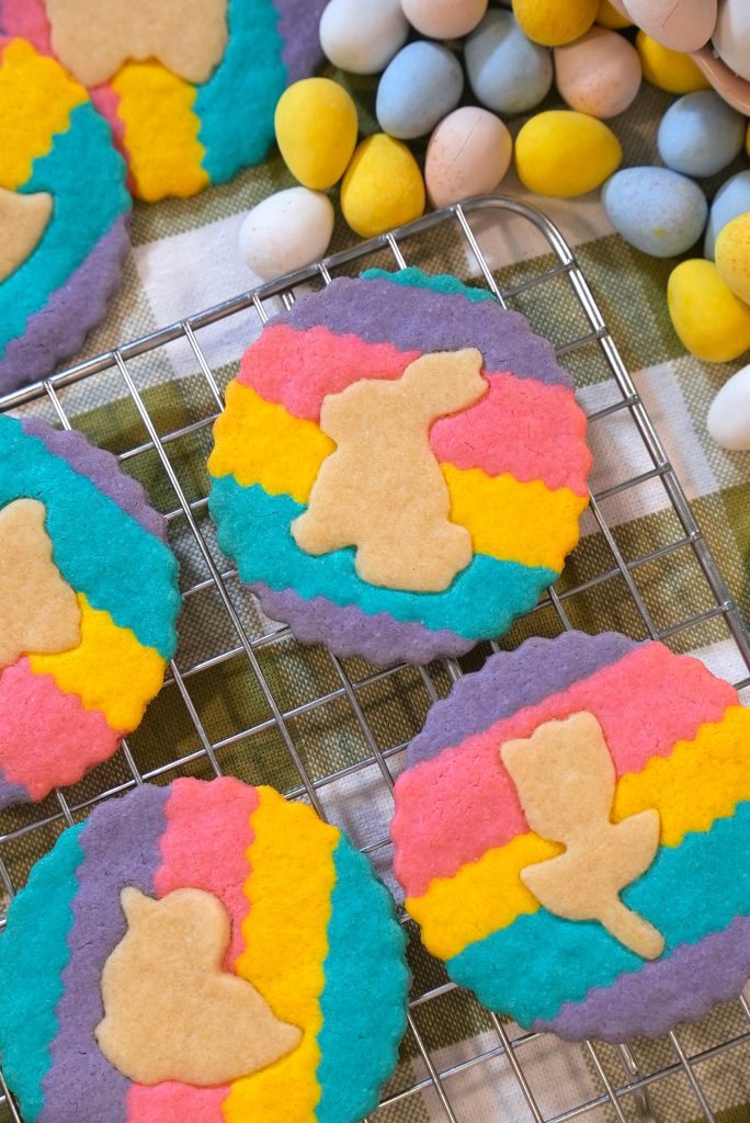 Colorful round cookies with zigzag stripes in blue, pink, and yellow are topped with tan Easter-themed shapes, including bunnies and chicks. Candy eggs are scattered nearby on a plaid cloth. Cookies cool on a wire rack.