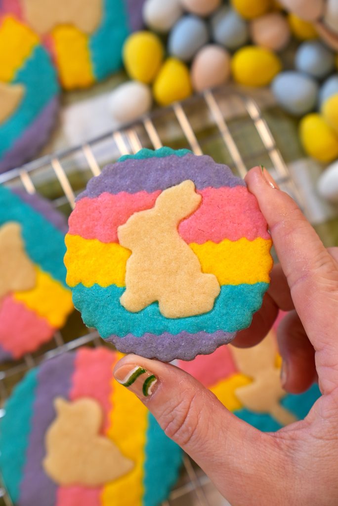 A hand holds a round, colorful cookie with wavy stripes of pink, purple, yellow, and blue and a bunny silhouette in the center. Other similar cookies and pastel candy eggs are in the background.