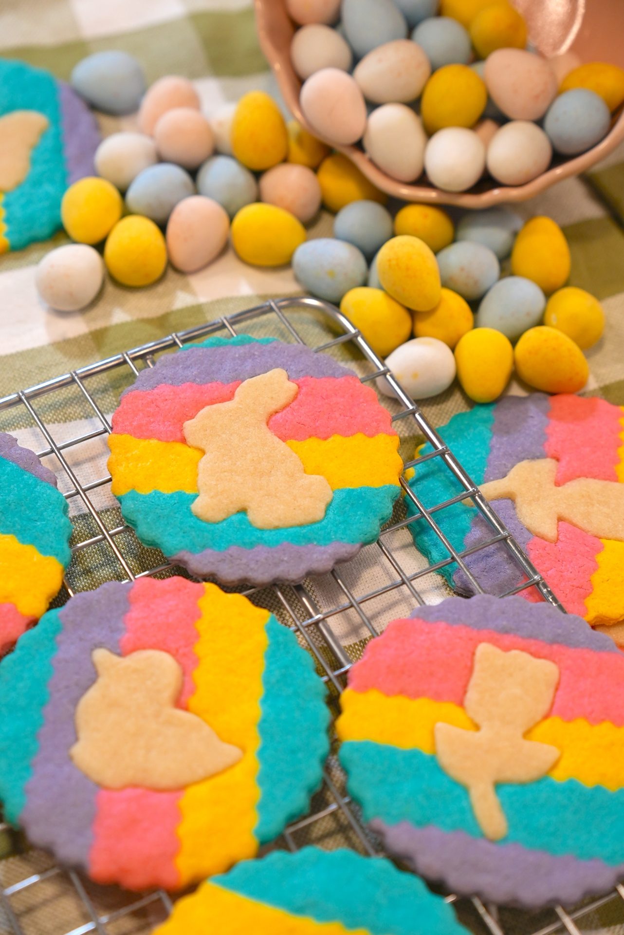 Colorful striped cookies with bunny and chick shapes are cooling on a wire rack. Behind them, pastel candy-coated chocolate eggs are scattered on a green and white checked cloth.
