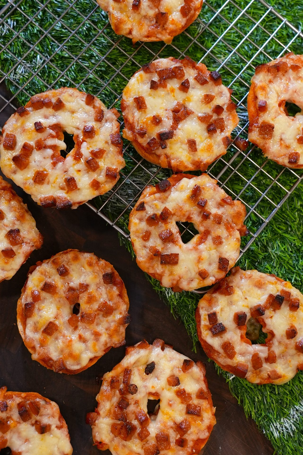 Several mini bagels topped with melted cheese and diced pepperoni are cooling on a wire rack, placed over a green grass-like surface. Some bagels are on the rack, while others are on the surface nearby.