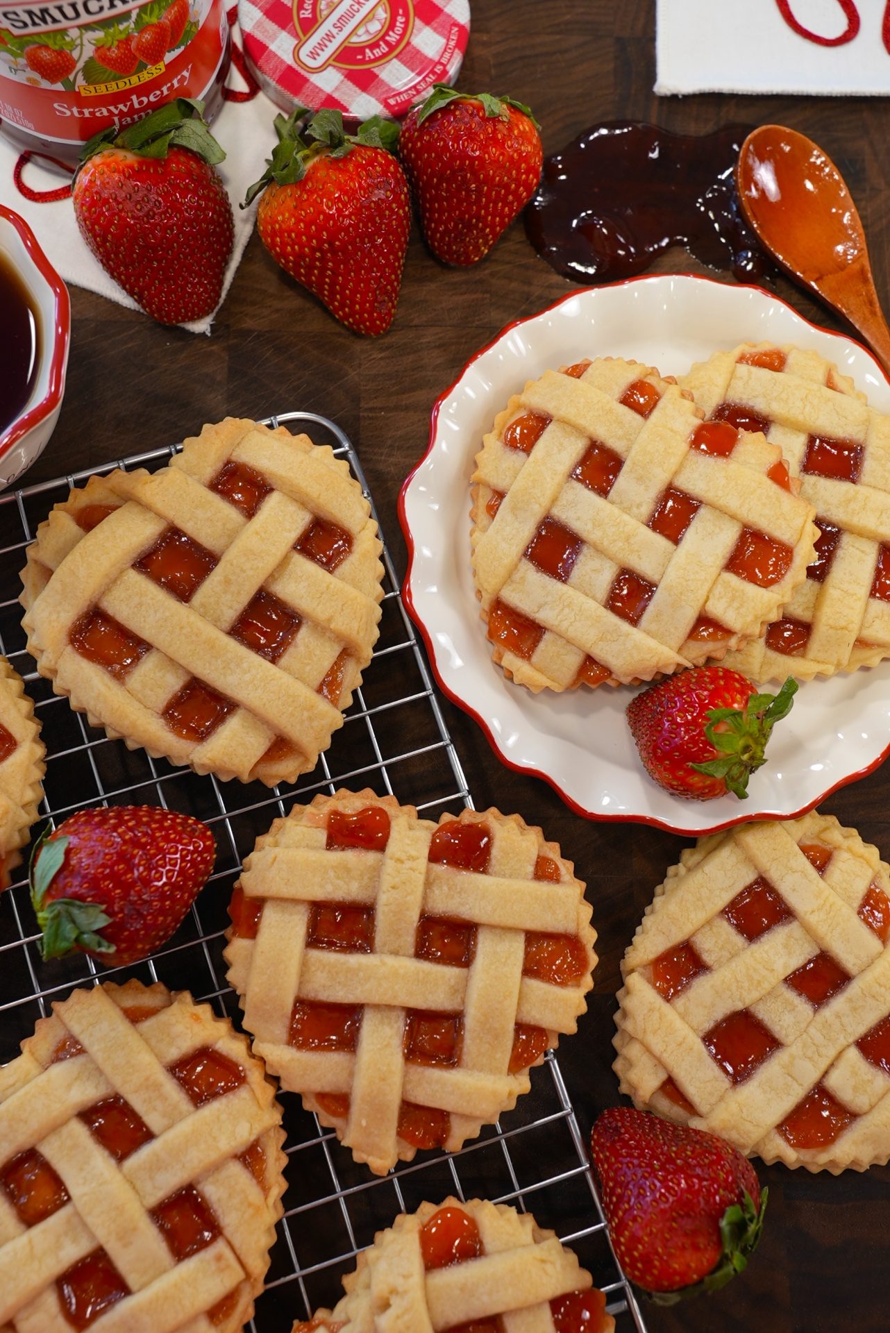 Heart-shaped strawberry jam tarts with lattice crusts are arranged on a cooling rack and plate, surrounded by fresh strawberries, jars of jam, and a spoon with strawberry jam on a dark wooden surface.