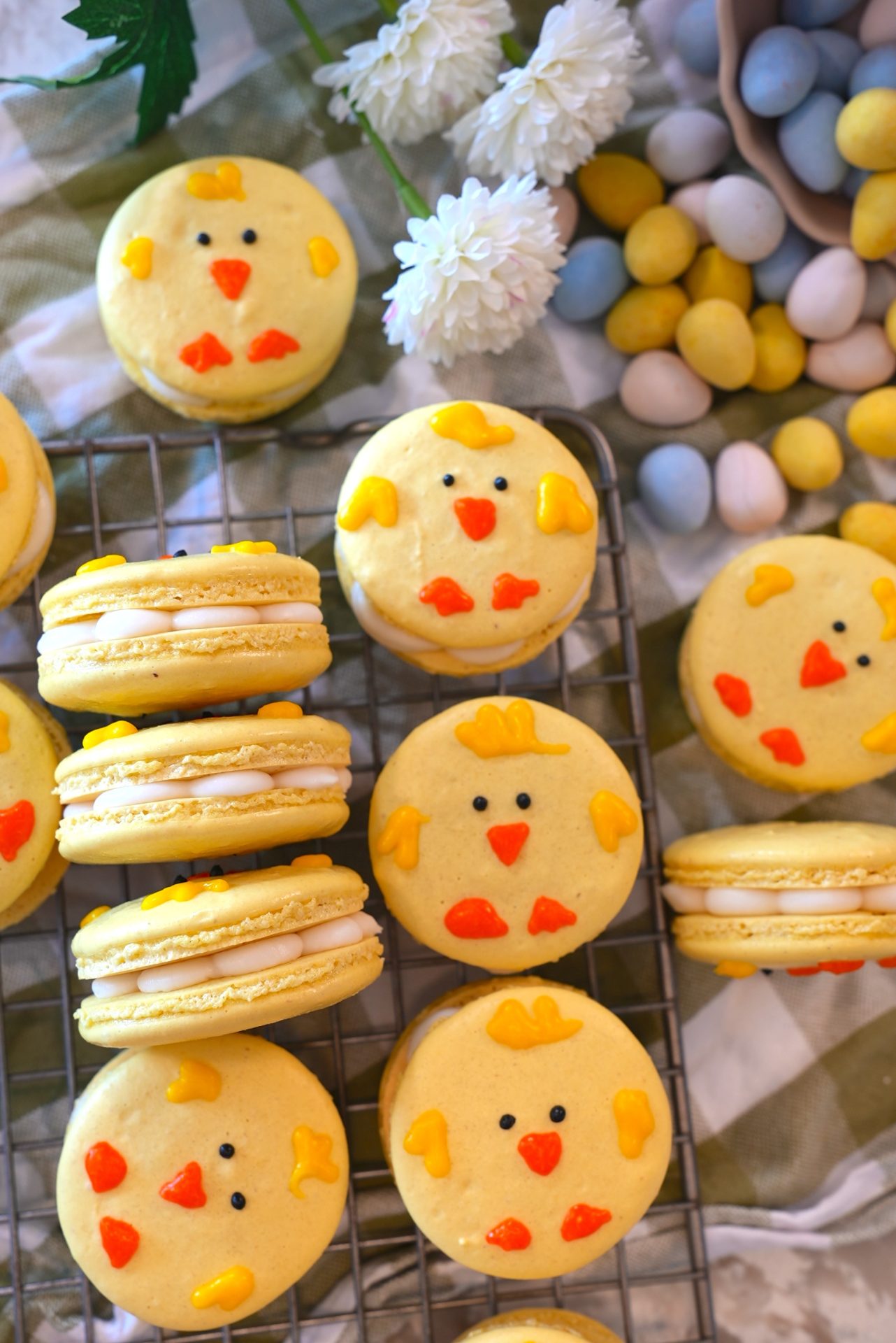Yellow chick-themed macarons with orange beaks and cheeks are arranged on a cooling rack and a checkered cloth, surrounded by white flowers and pastel candy eggs.