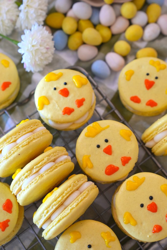 Yellow macarons decorated as chick faces with orange and yellow icing are displayed on a cooling rack, surrounded by pastel-colored mini eggs and white flowers.